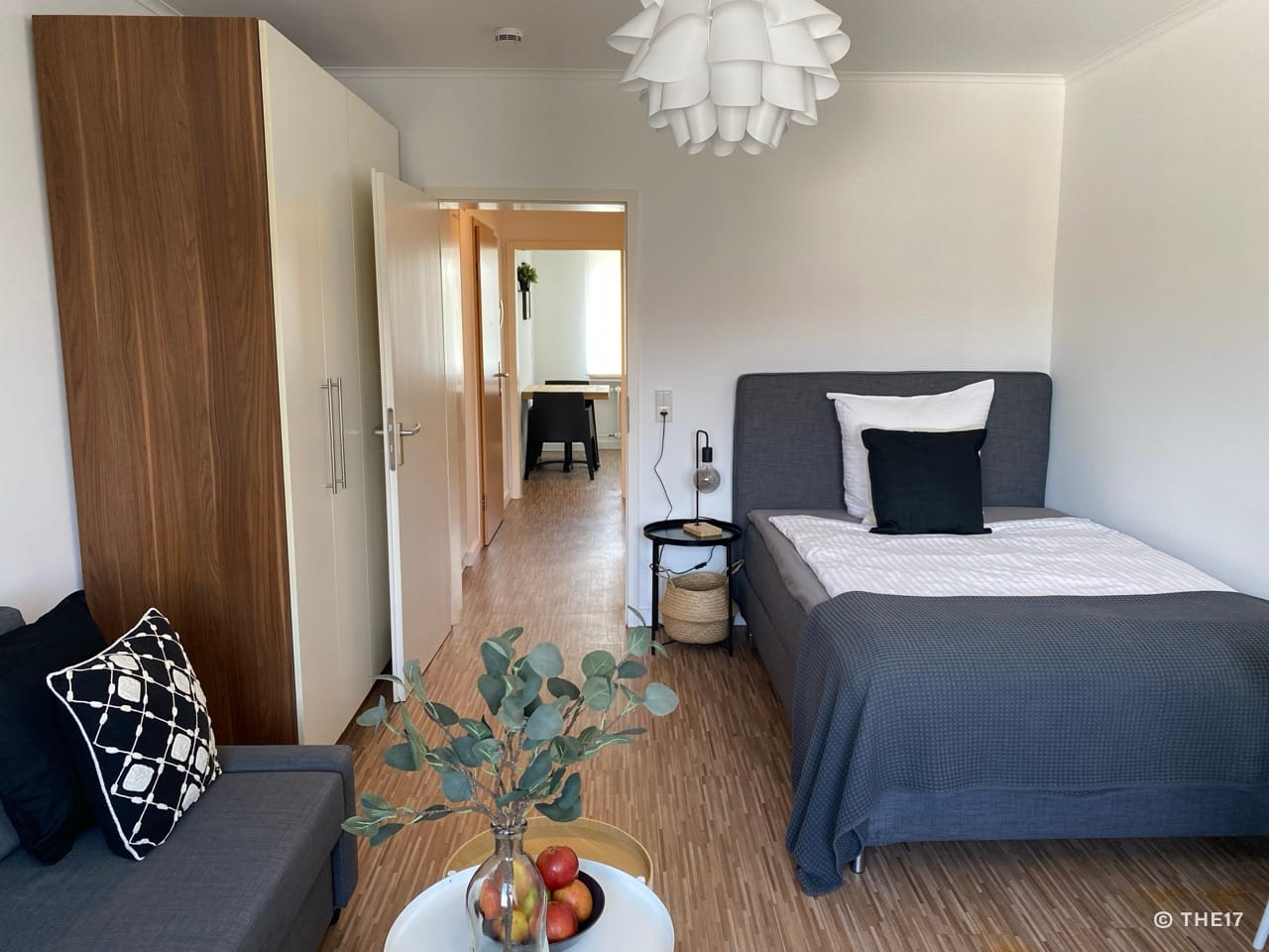 Schlafzimmer mit grauem Bett, schwarzem Nachttisch und Sofa, Holzkleiderschrank und Pflanze in Vase auf weißem Tisch.