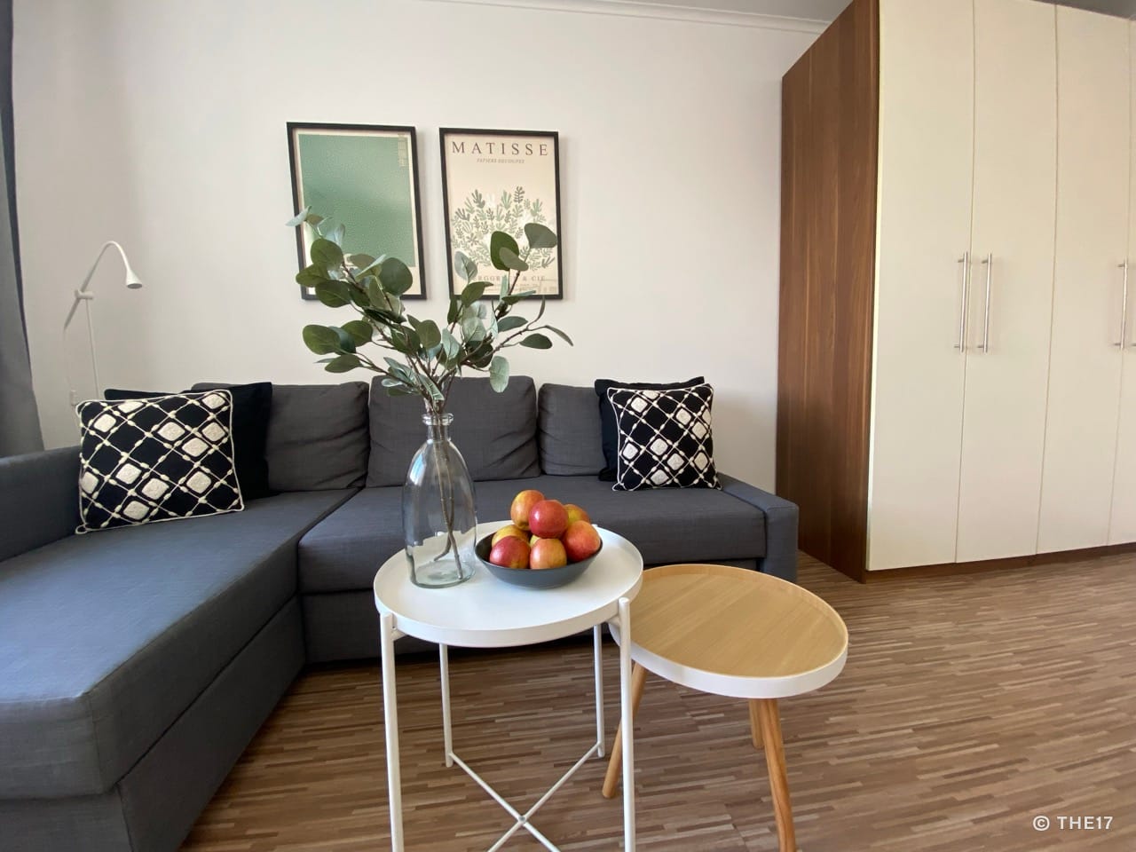 Wohnzimmer mit grau-lila Ecksofa, zwei kleinen Tischen, einer mit Äpfeln, Pflanzen in Vase, zwei Wandbildern und großem Schrank.