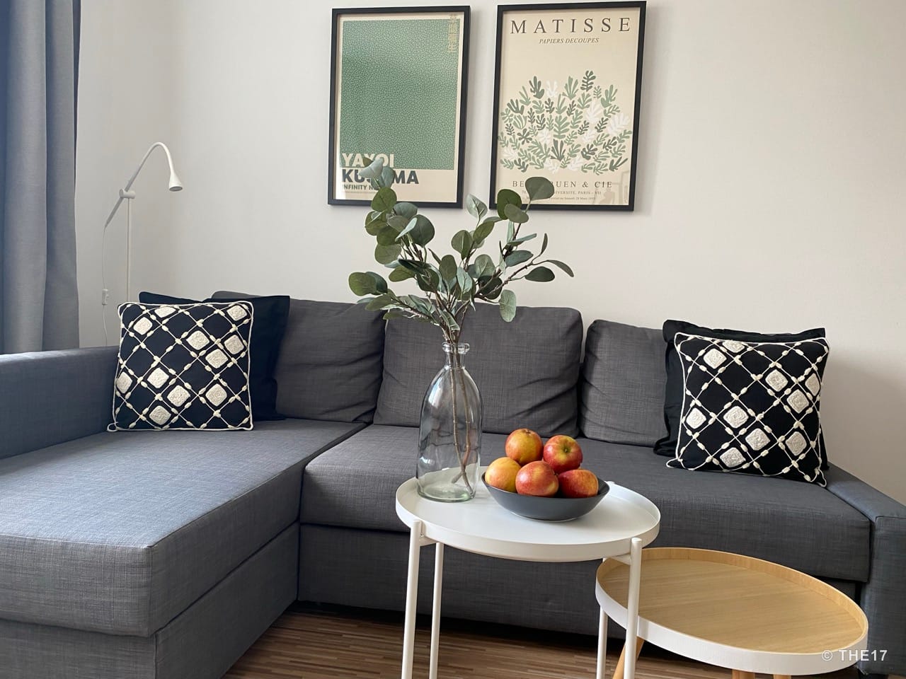 Gray L-shaped sofa with black and white pillows, two framed artworks on the wall, table with glass vase and apples.