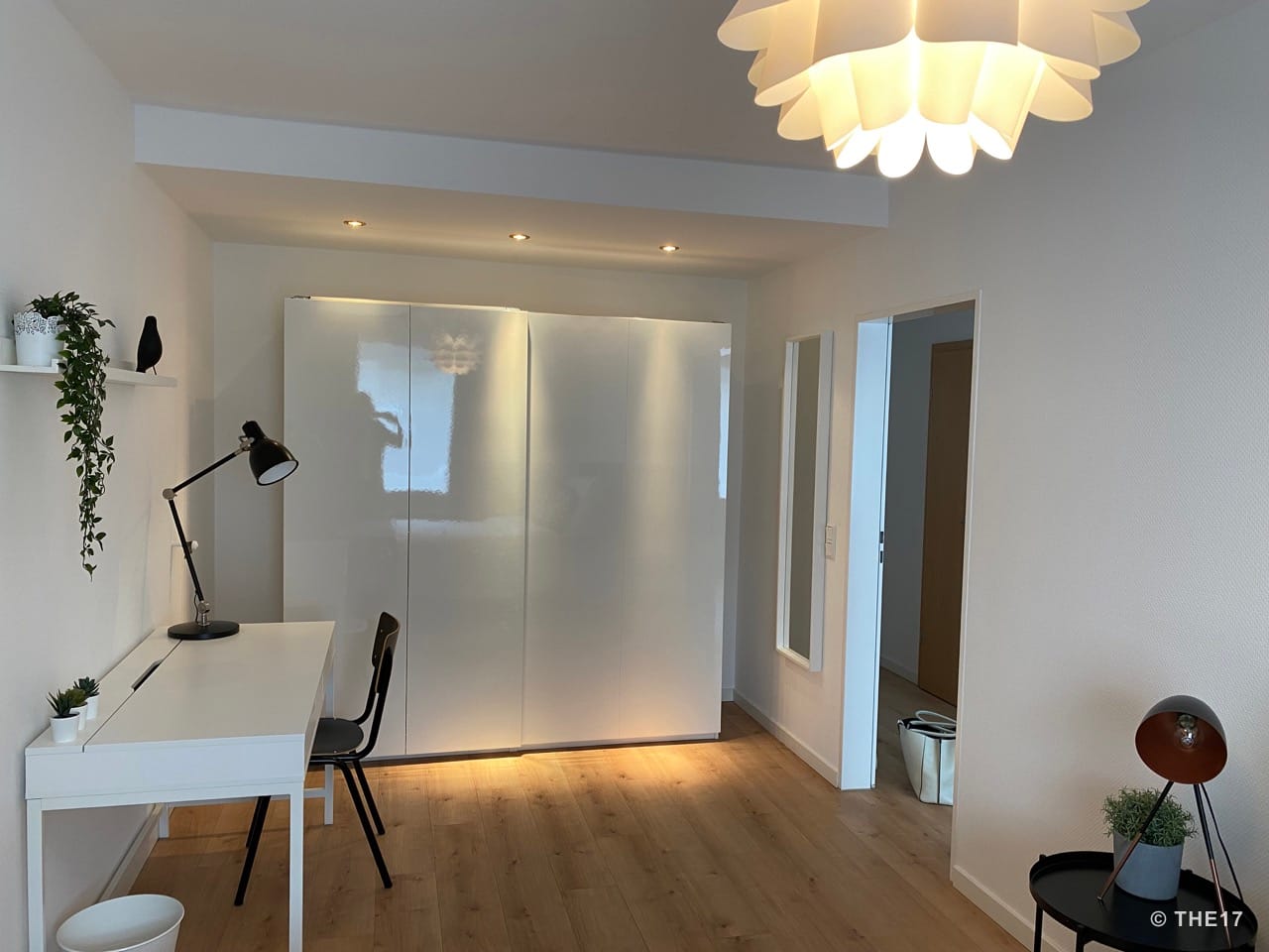 Bright room with white desk, black chair, large white wardrobe, wooden floor, and modern lighting.