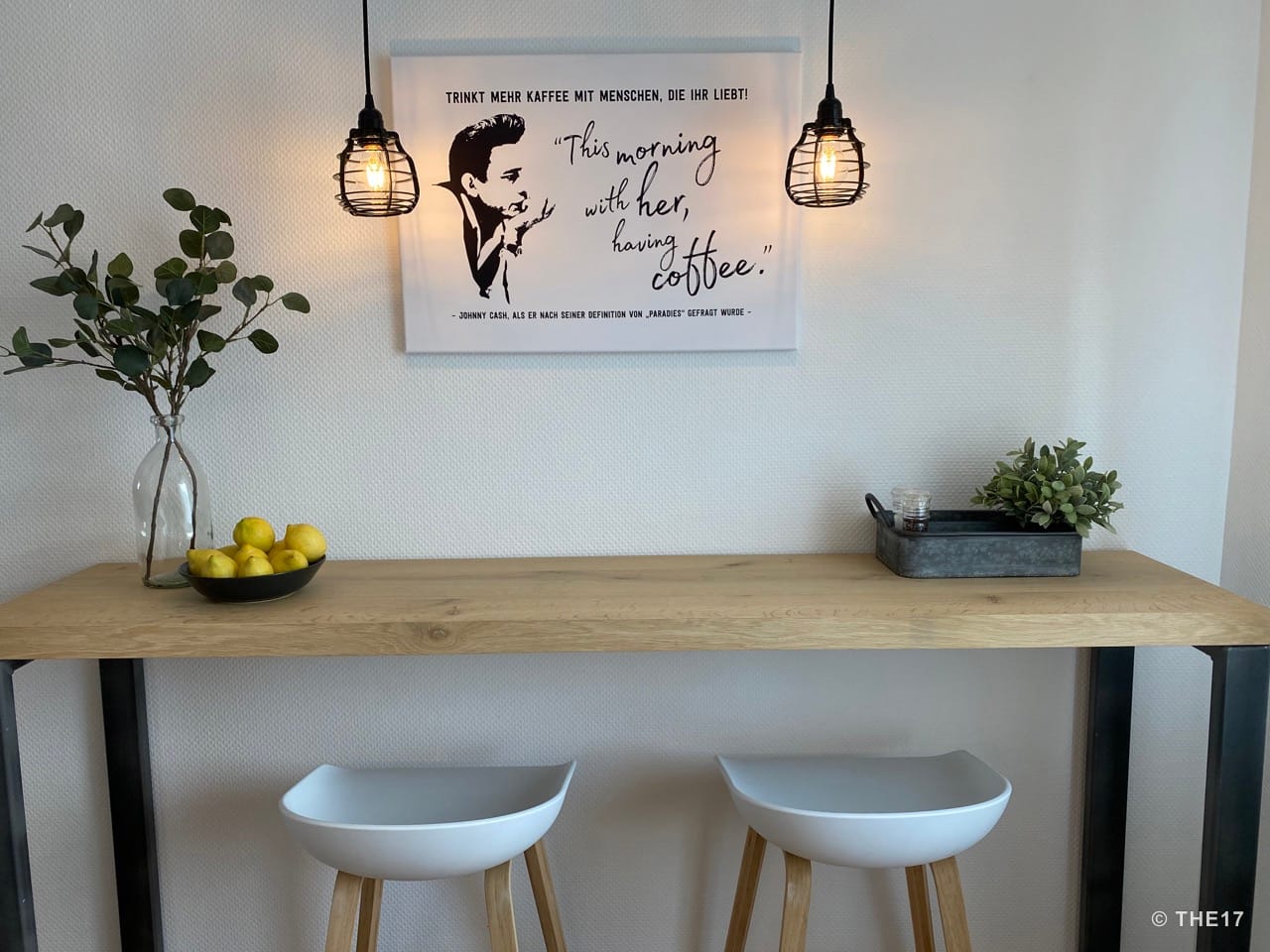 Wooden kitchen table with two white stools, hanging lamps, bowl of lemons, and green plant in glass vase.