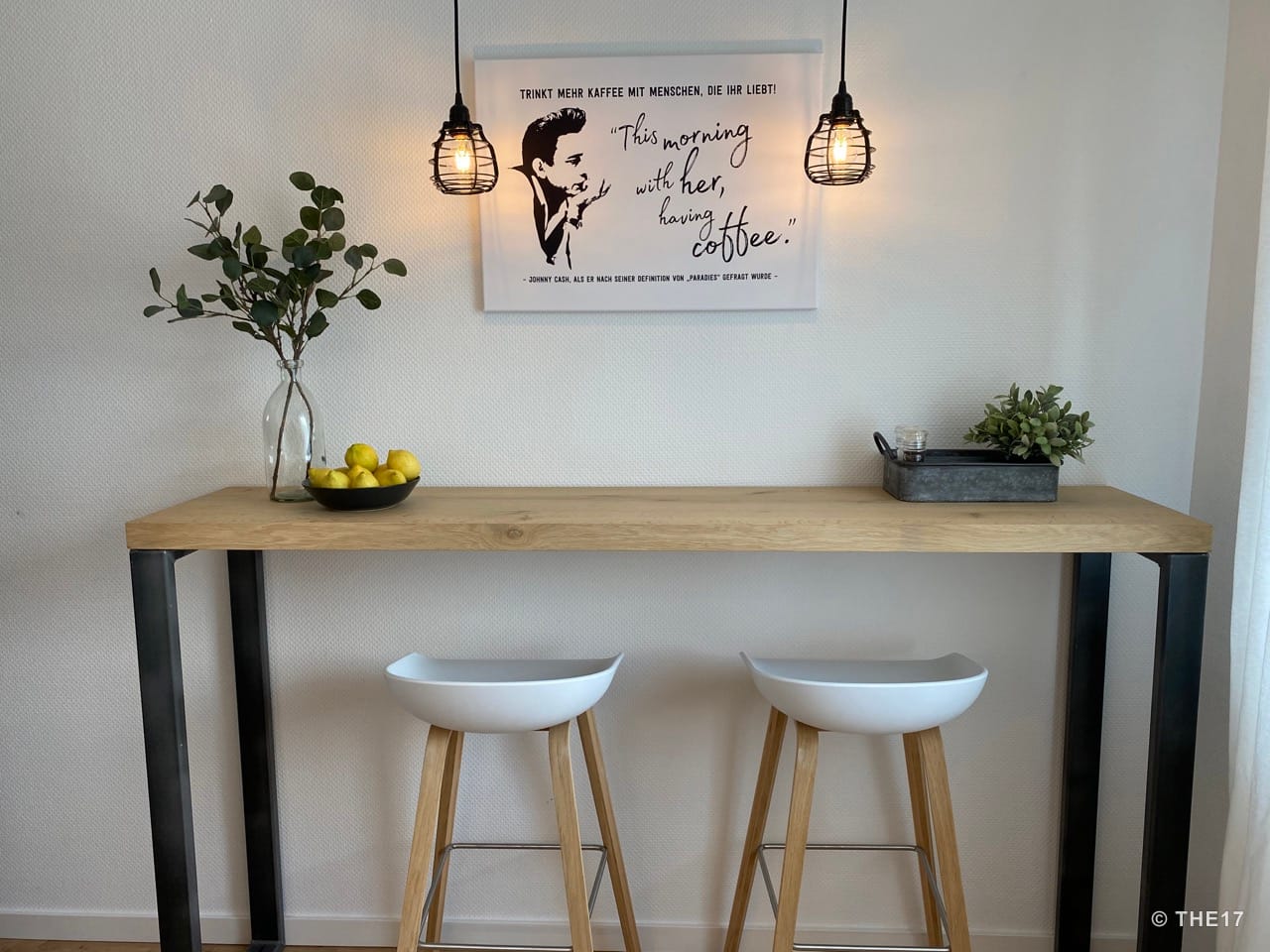 Tall wooden table with black metal legs, two white stools, hanging lamps, and a wall art featuring a coffee quote.