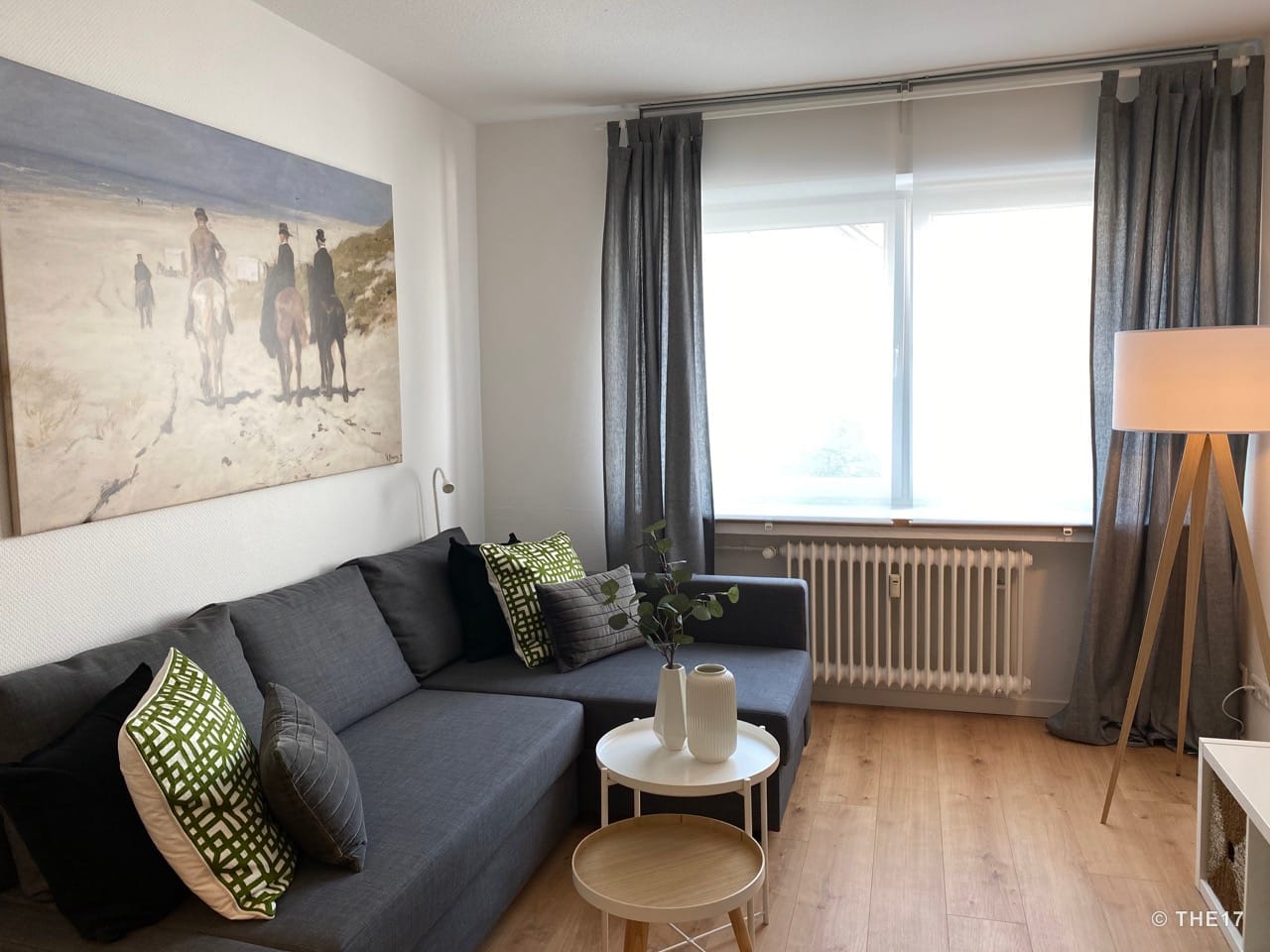 Living room with gray sectional sofa, green and gray pillows, two side tables, floor lamp, and large window with gray curtains.