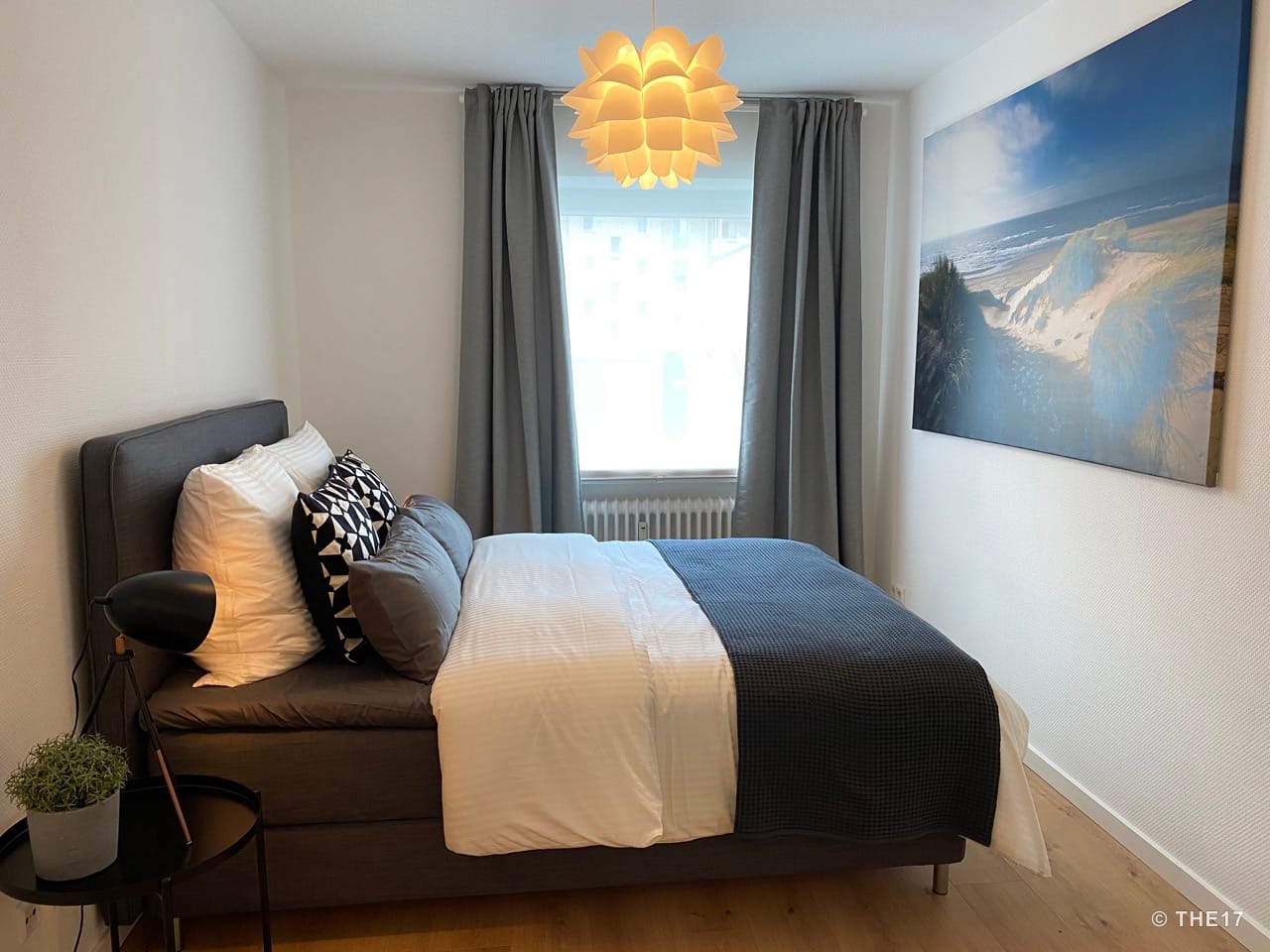 Bedroom with double bed, gray headboard, pillows, bedside table with lamp and plant, and a beach picture on the wall.