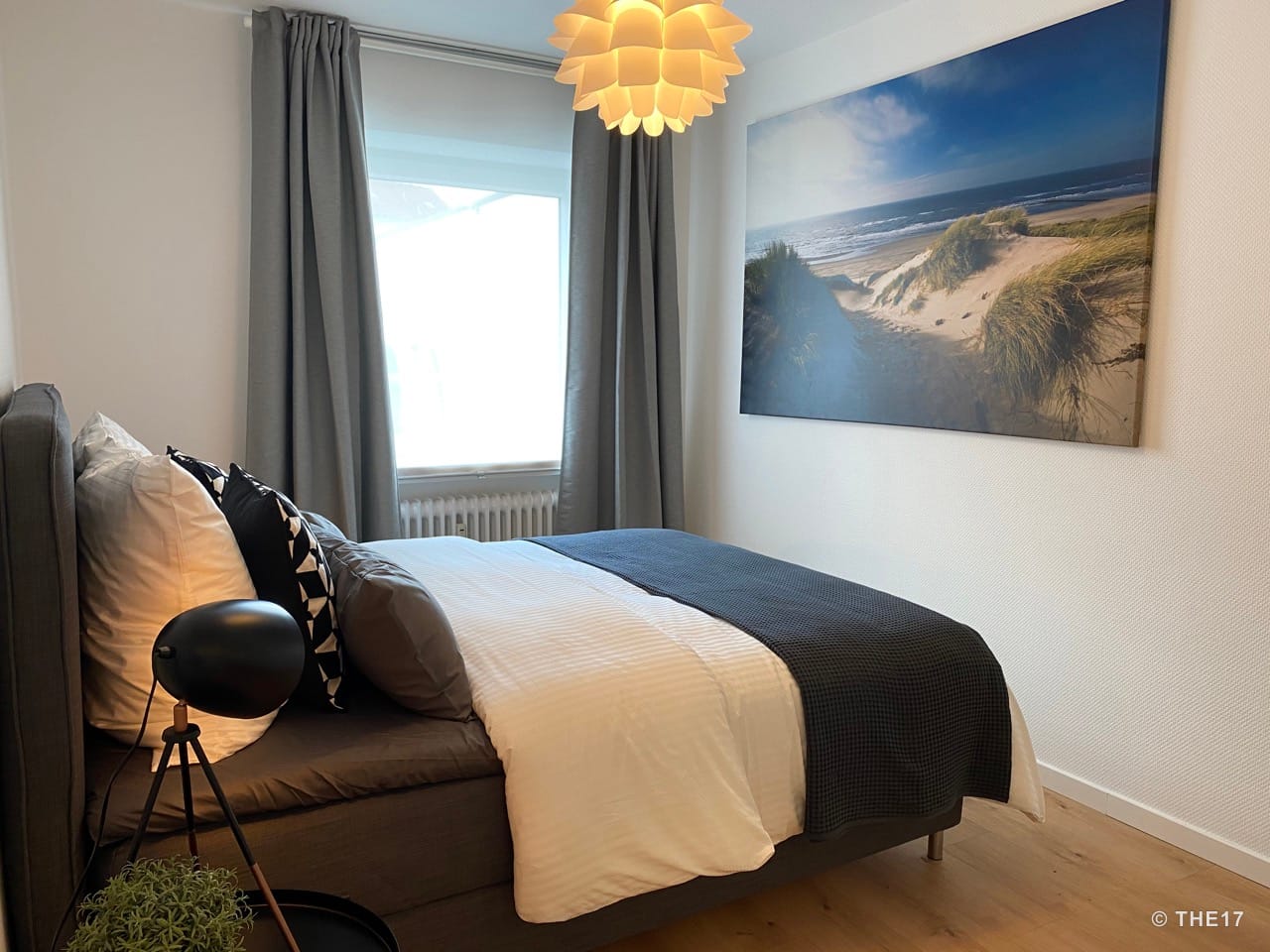Bedroom with gray bed, white and black pillows, large window with gray curtains, dune beach artwork, and wooden floor.