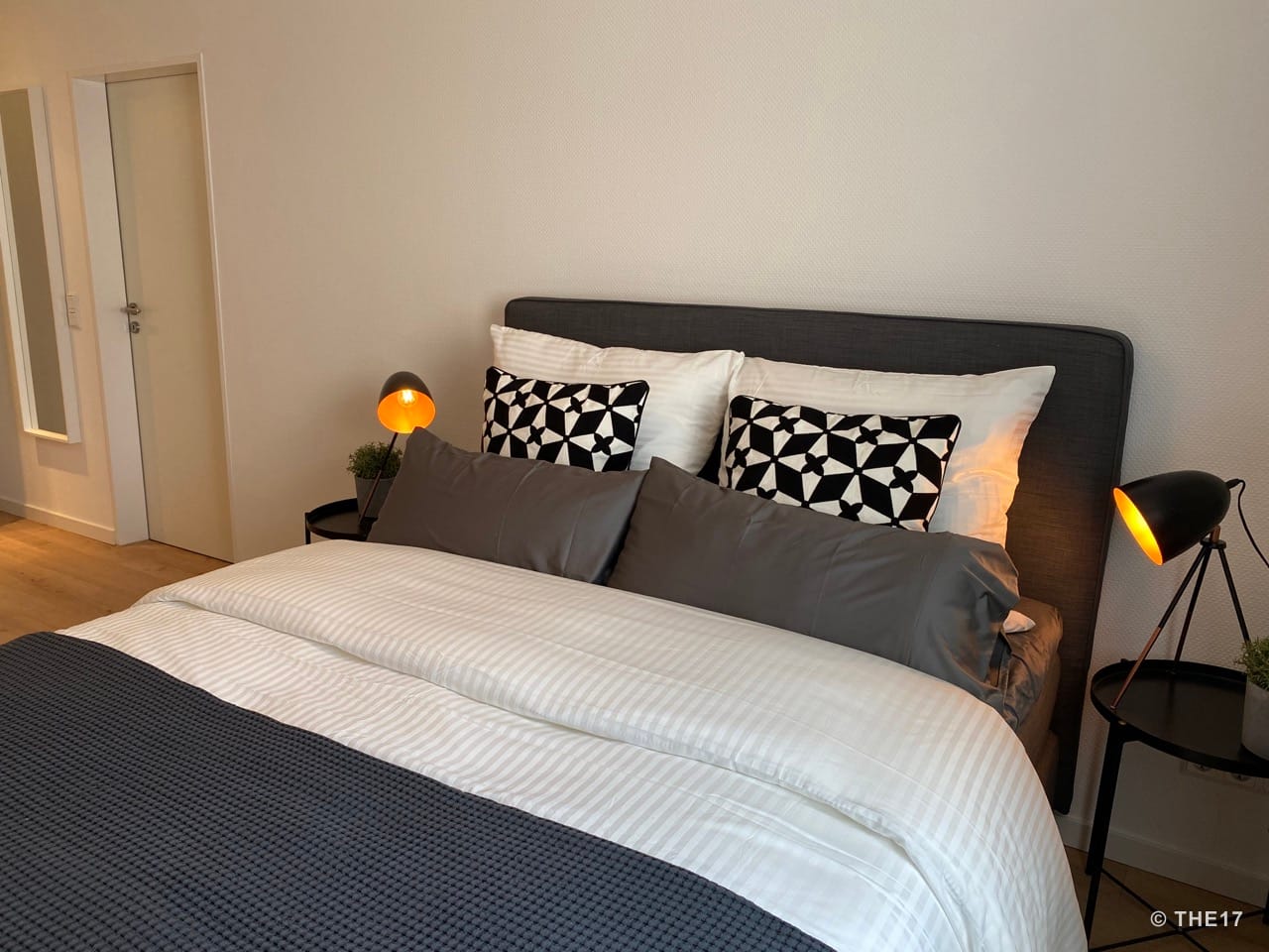 Bedroom with gray headboard, white and patterned pillows, gray bedding, and two black bedside lamps.