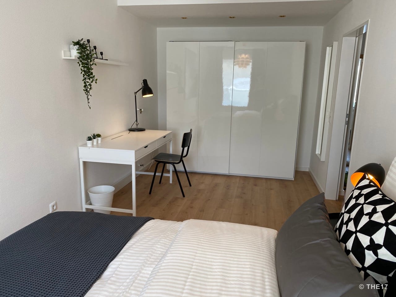 Bedroom with white desk, black chair, large white wardrobe, and cozy bed with patterned pillows.