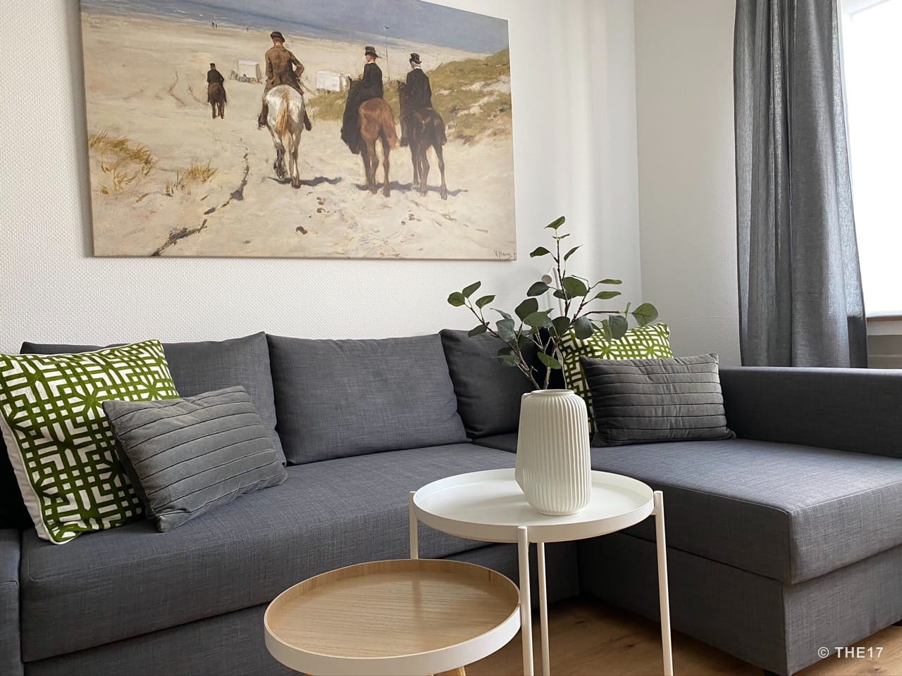 Graues Ecksofa mit zwei grünen und zwei grauen Kissen, weiße Beistelltische mit weißer Vase und Pflanzenzweigen, Gemälde an weißer Wand.