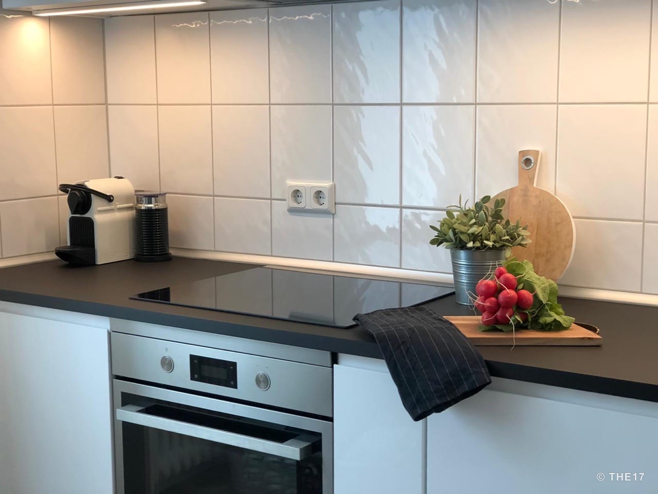 Modern kitchen with black countertops, built-in oven, coffee machine, cutting board, and radishes on countertop.
