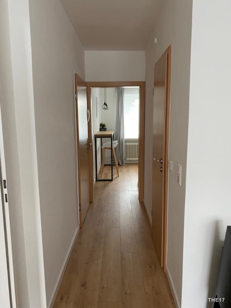 Hallway with wooden floor, two wooden doors on the right and left, view into a bright room with a bar table and stool.