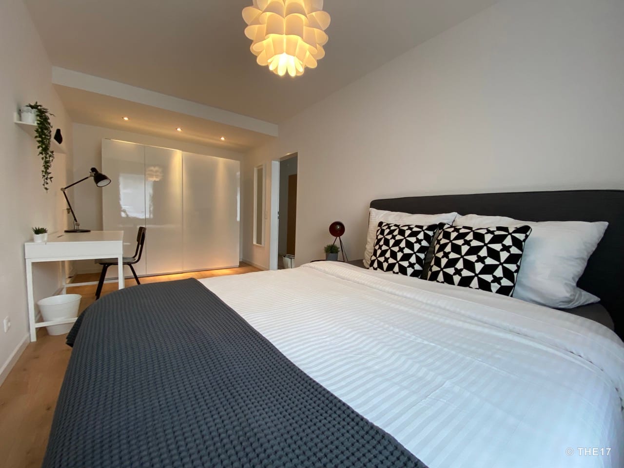 Bedroom with large bed, white bedding, black patterned pillows, desk, chair, and white wardrobe with lights.