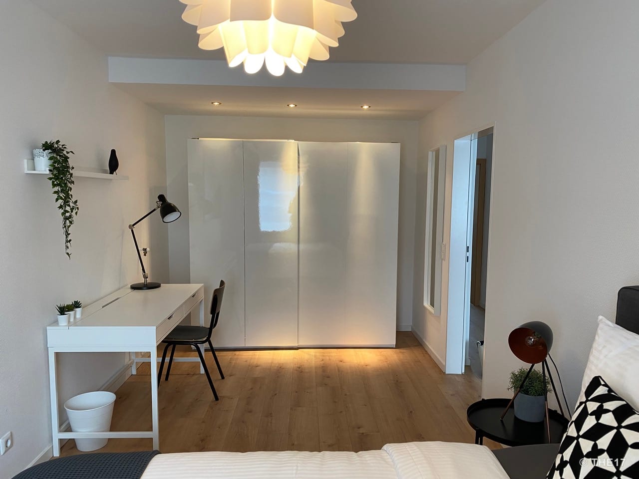 Bedroom with white desk, black chair, small wall shelf, large white wardrobe, and wooden flooring.
