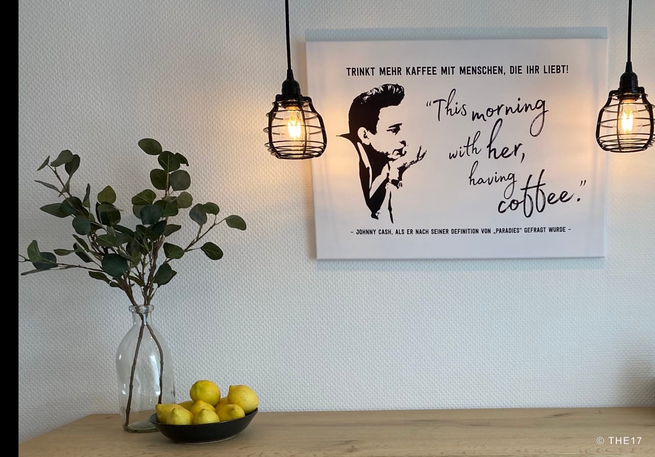 Wall sign with Johnny Cash quote, two hanging wire lamps lit, vase with branches and bowl with lemons on wooden table.