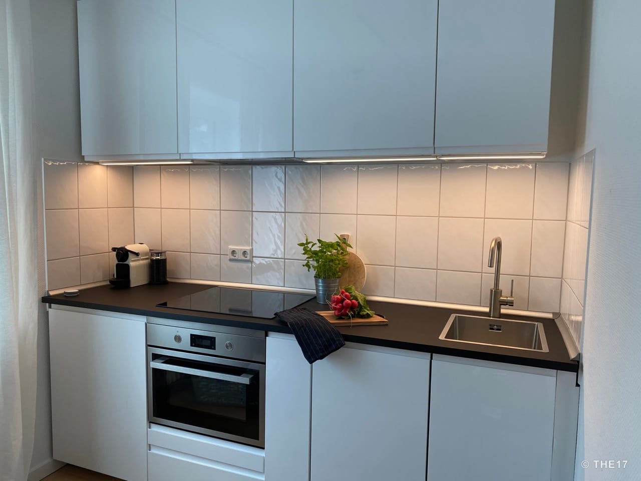 Modern white kitchen unit with oven, ceramic hob, sink, coffee machine, and fresh radishes on black countertop.