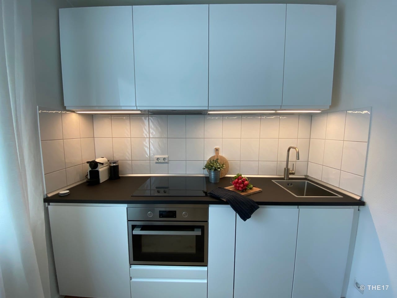 Modern kitchen with white cabinets, stovetop, oven, sink, coffee machine, and utensils on a black countertop under tiled backsplash.
