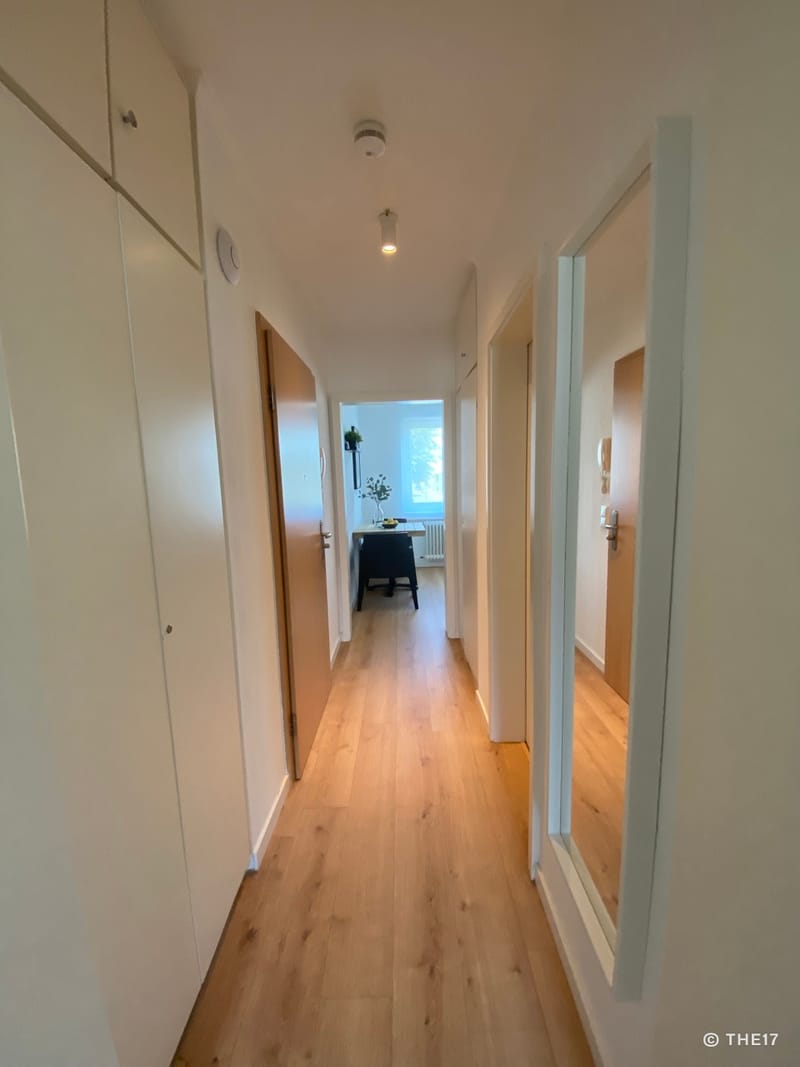 Hallway with light walls, wooden floor, white cabinets on left, large wall mirror on right, view into a bright room ahead.