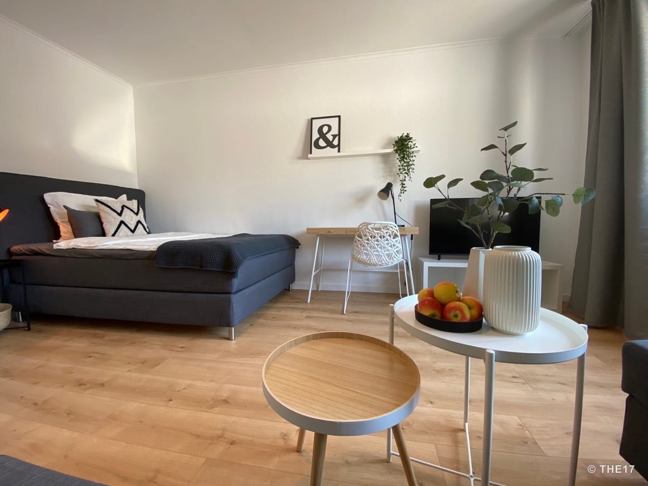 Bedroom with gray bed, wooden floor, desk, TV, white tables with apples, and green plant arrangement.