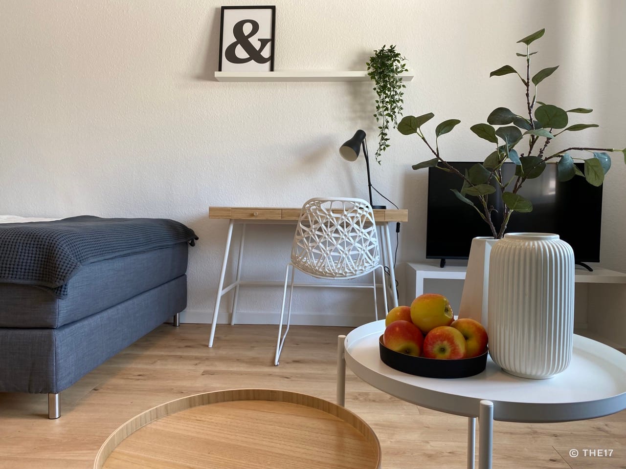 Room with gray bed, wooden desk with white chair, wall shelf with decoration, round tables with apples and plant.
