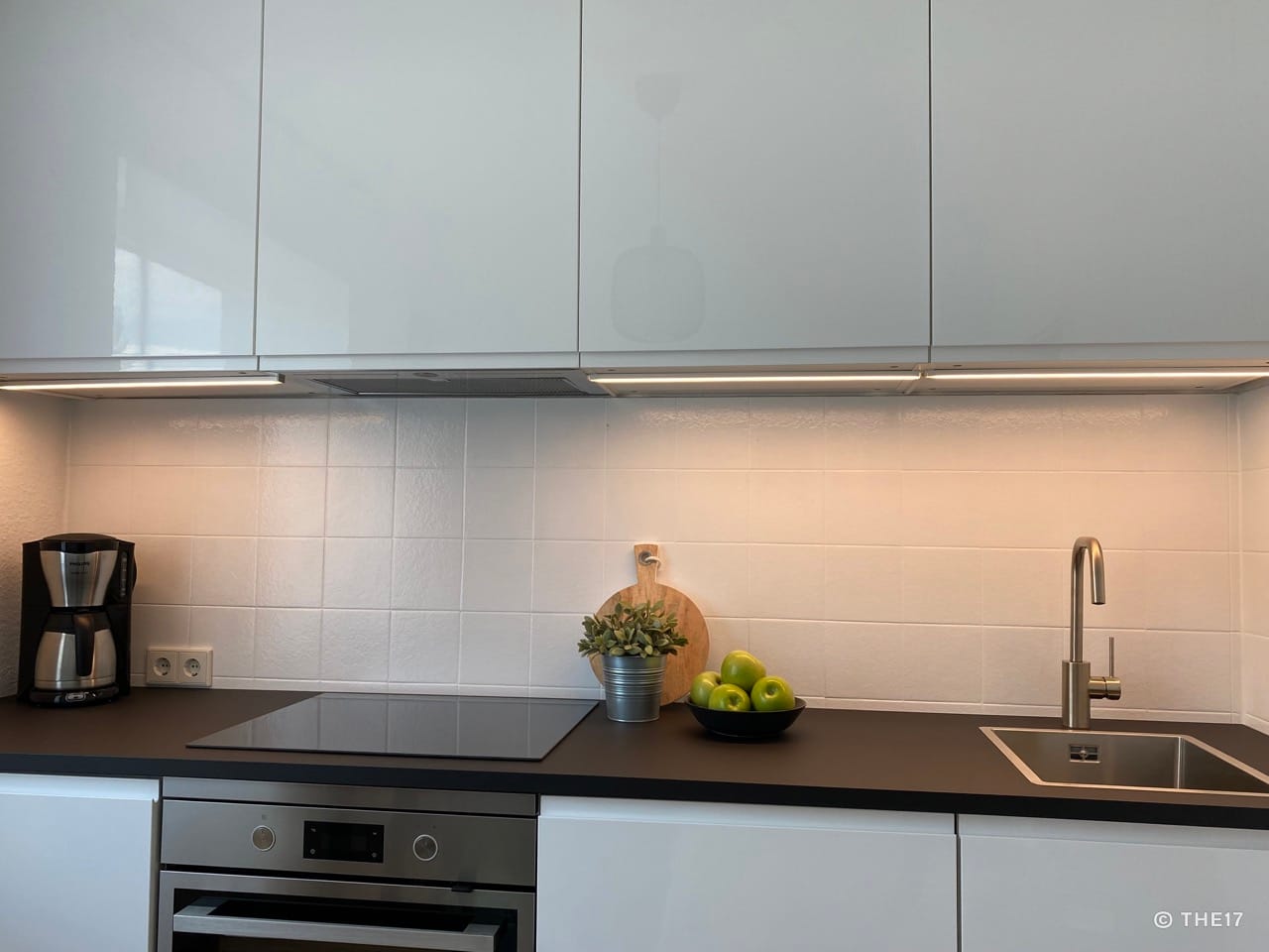Modern kitchen area with black countertop, induction hob, sink, coffee maker, and white cabinets.