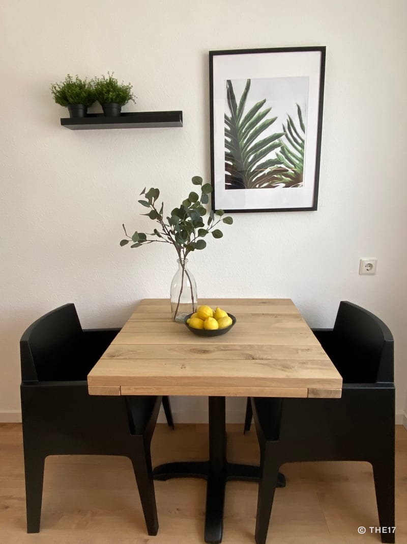 Wooden table with two black chairs, vase with green branches, bowl of lemons, plants on black wall shelf, leaf print on white wall.