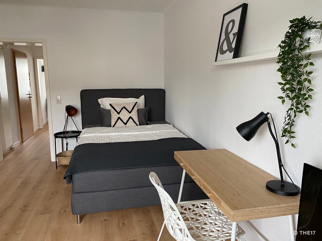 Bedroom with gray bed, wooden floor, desk with black lamp, white chair, and wall-mounted leafy plant.