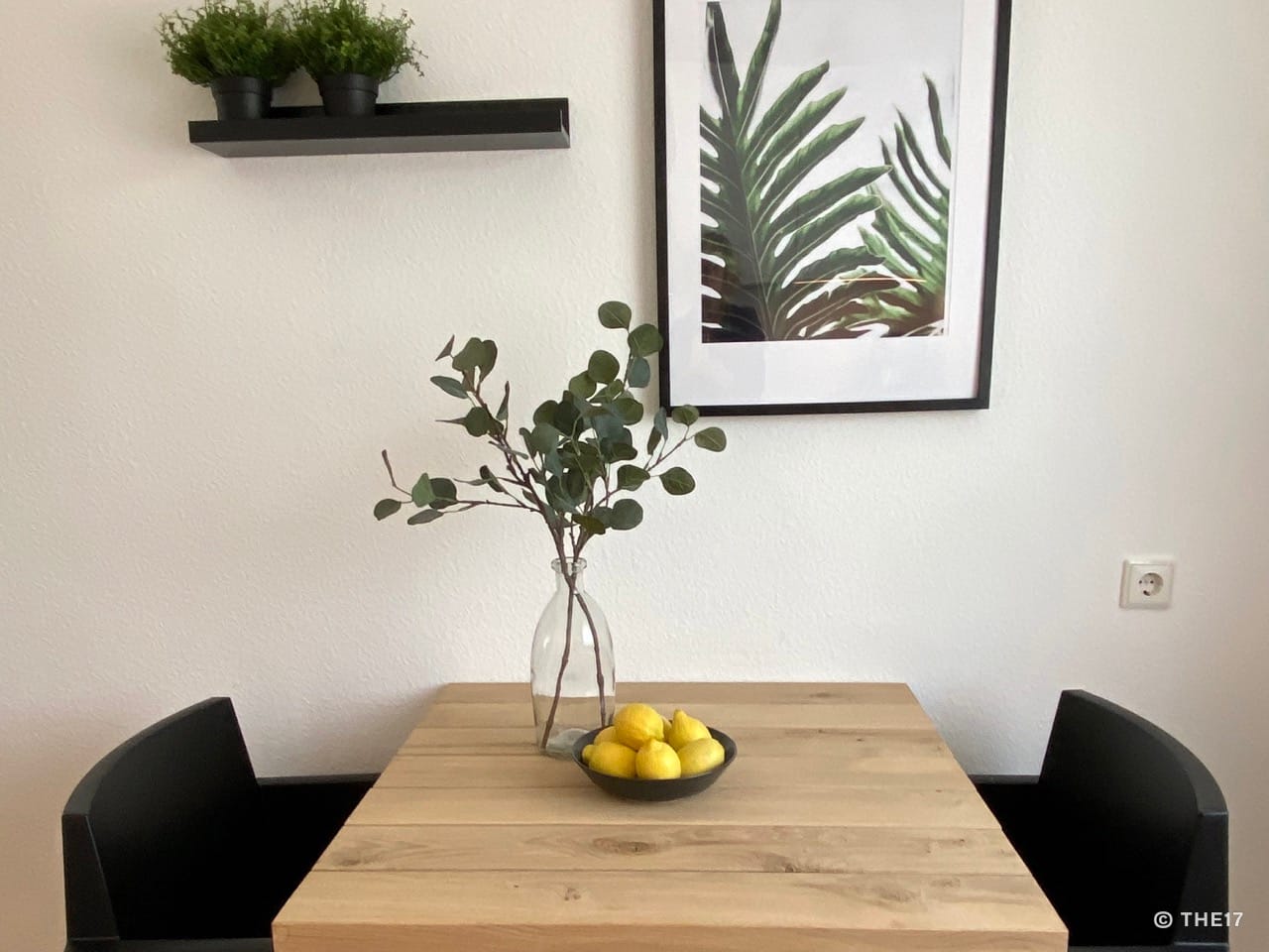 Wooden table with black chairs on both sides, a glass vase with branches and a bowl of lemons on the table.