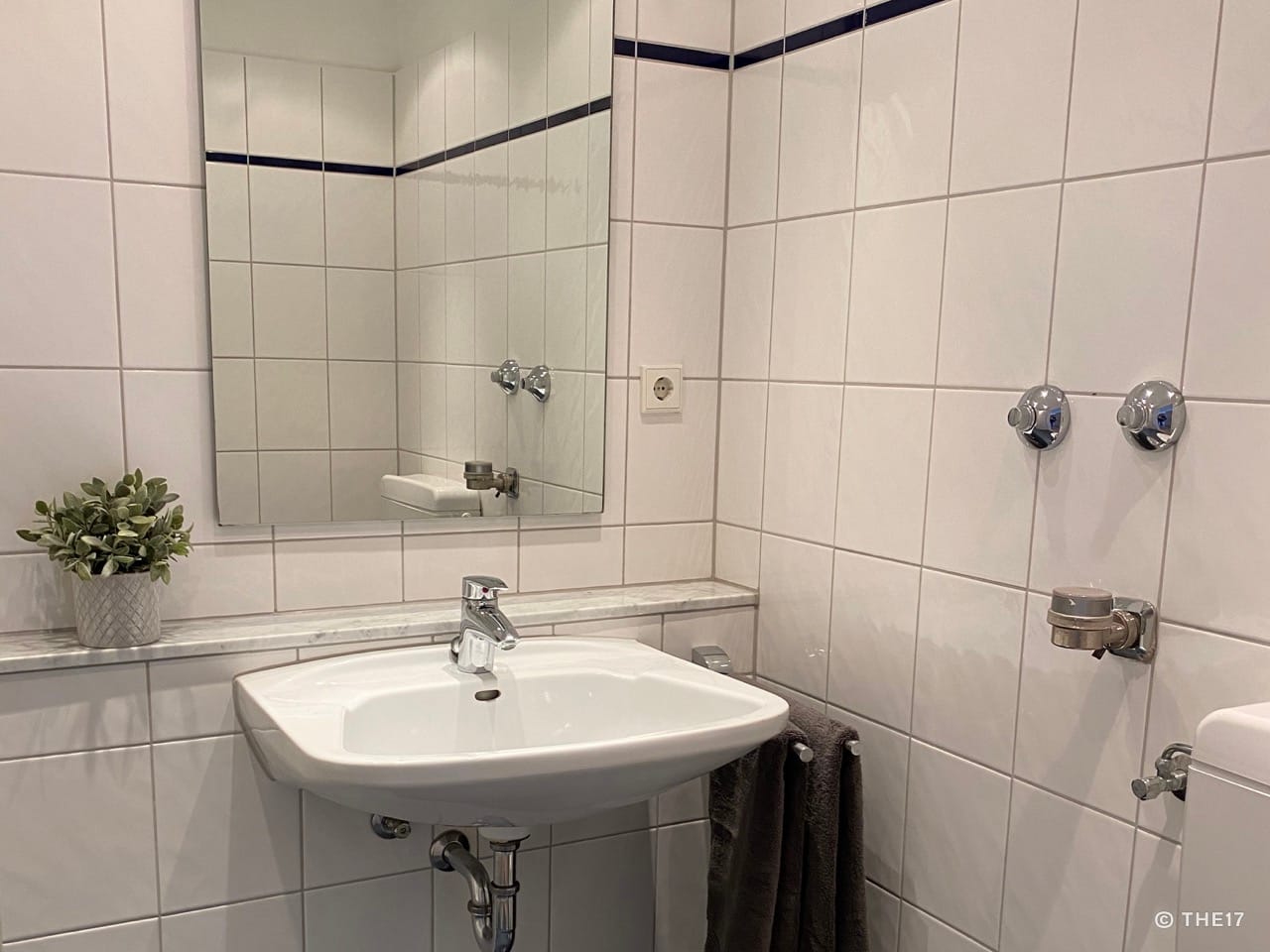 Small bathroom with white tiled sink, mirror, gray towel, and potted plant on the ledge.