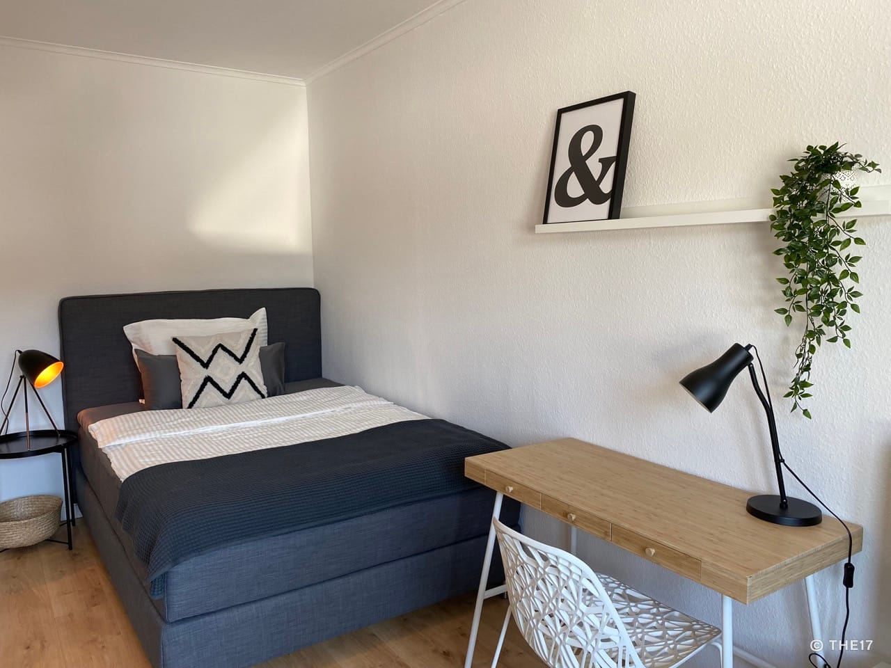 Bedroom with gray bed, decorative pillows, wooden desk, white chair, wall shelf with framed print and plant.