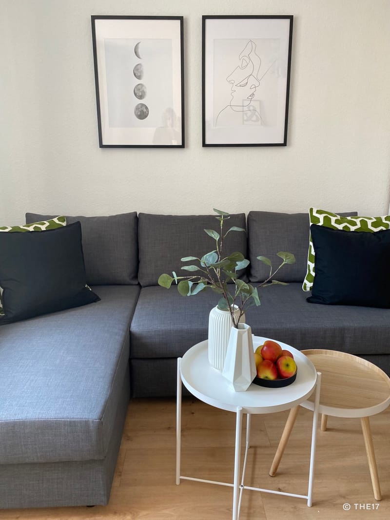 Gray sectional sofa with black and green pillows under two framed black and white pictures on a white wall.