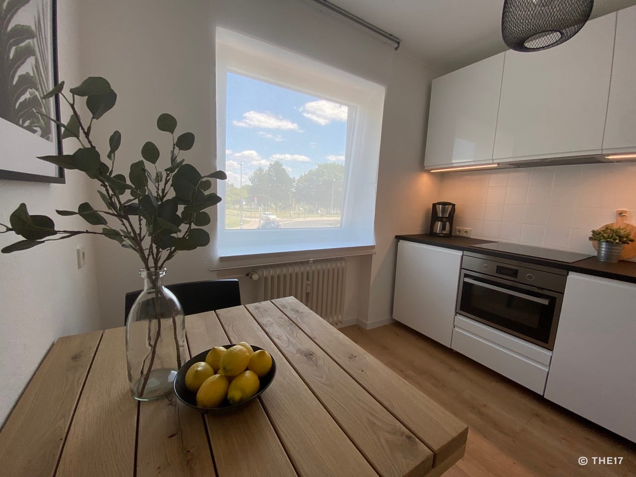 Small kitchen with wooden table, bowl of lemons, window shade, white cabinets, and built-in oven.