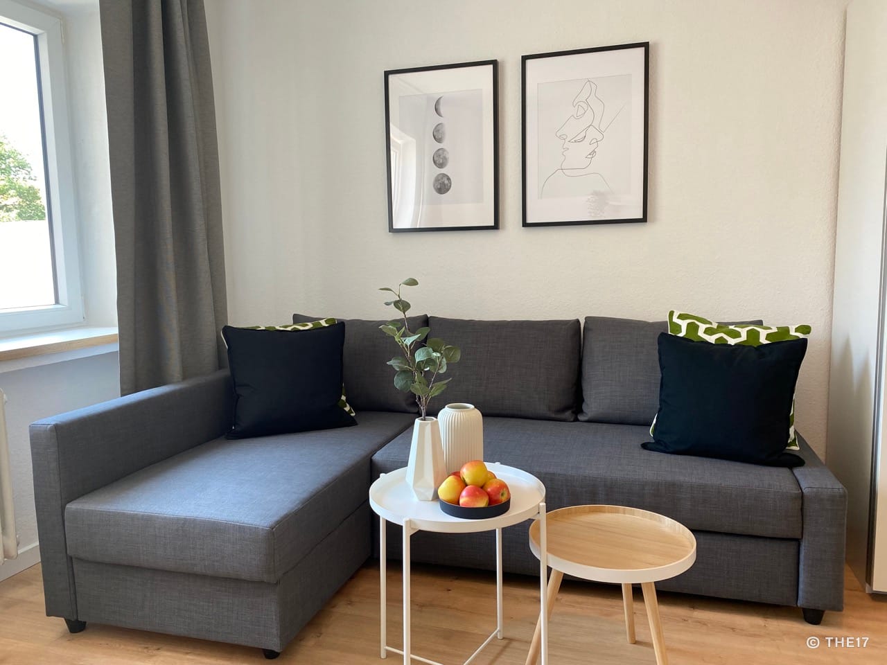 Gray sectional sofa with black and patterned pillows, two side tables with vases and apples, two framed black and white pictures on white wall.