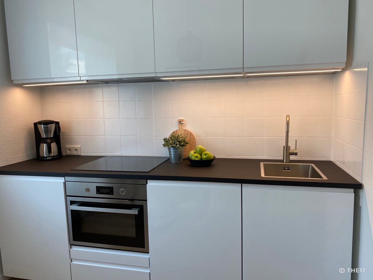 Modern kitchen with white cabinets, black countertop, oven, stovetop, coffee maker, and stainless steel sink.