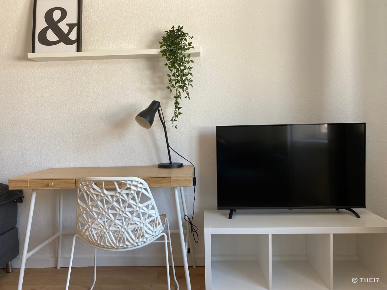 Modern workspace with wooden desk, white designer chair, black lamp, and TV on white shelf next to wall with potted plant.