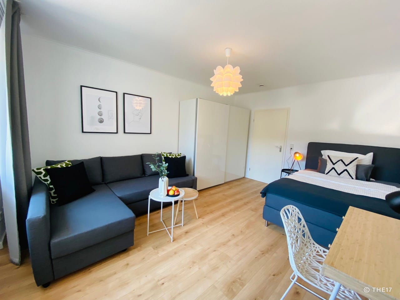 Bright room with grey corner sofa, two side tables, double bed, white wardrobe, and wooden floor.