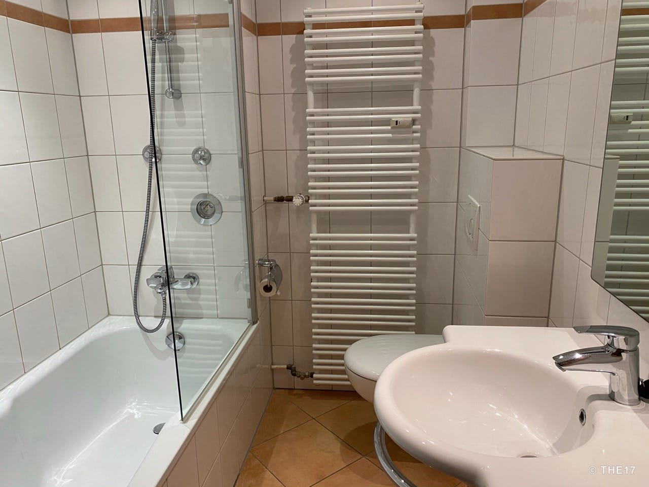 Kleines Badezimmer mit Badewanne, Glasdusche, weißer Heizung, Toilette und Waschbecken mit Chromarmatur.