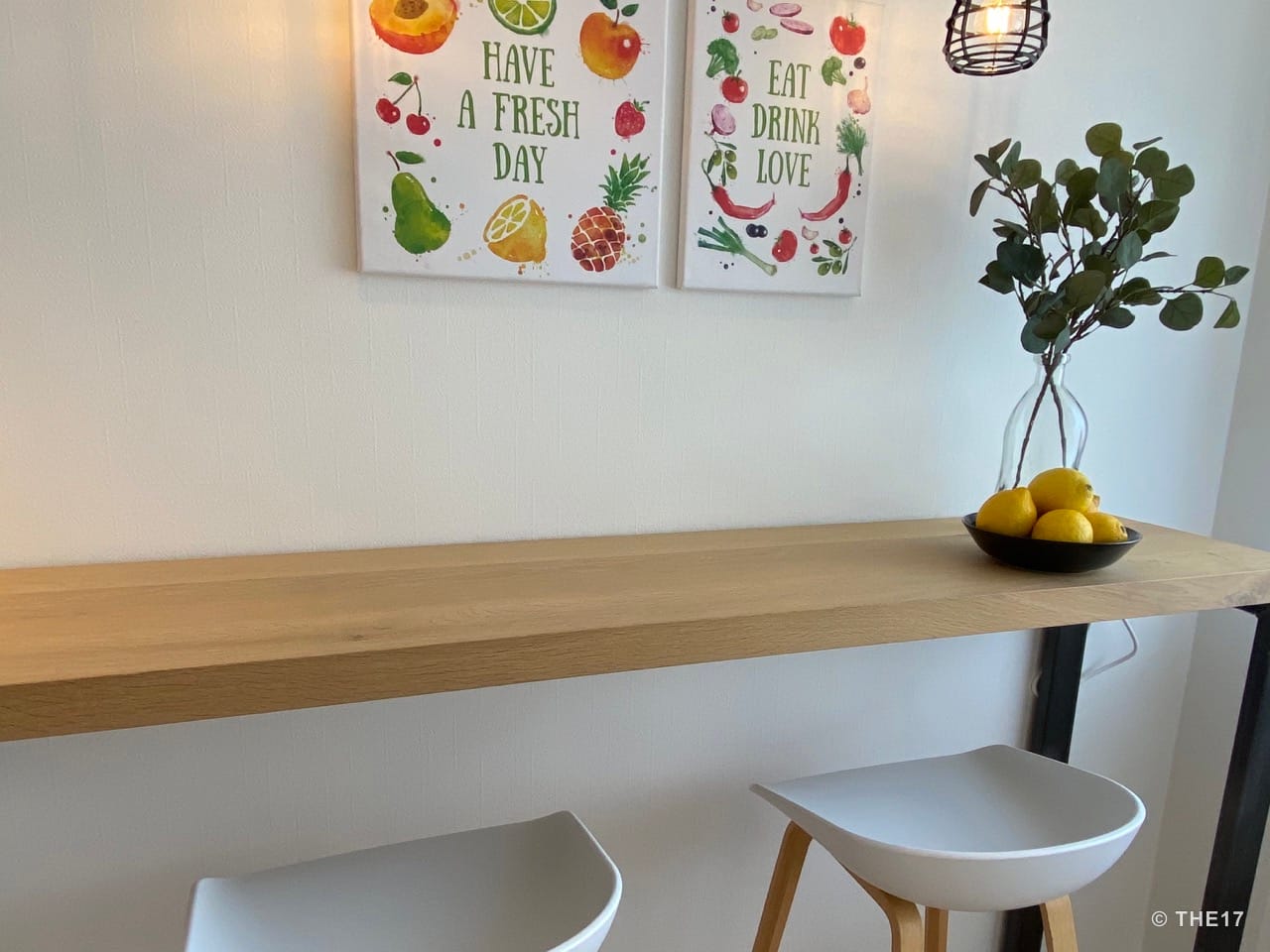 Küchenbereich mit Holztheke, zwei weißen Barhockern, Pflanzen in Vase und Wandbildern mit Obst und Sprüchen.