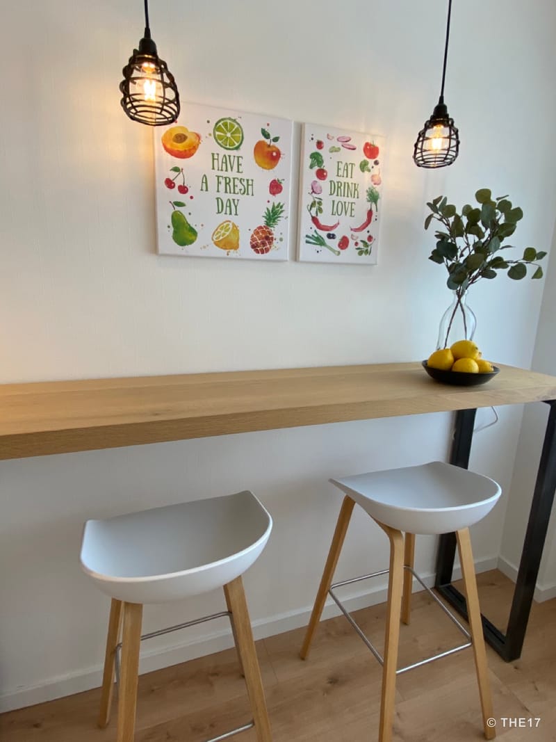 Küchenbar mit zwei weißen Hockern, Holzplatte, zwei Wandbildern mit Obstmotiven und hängenden Glühbirnen.