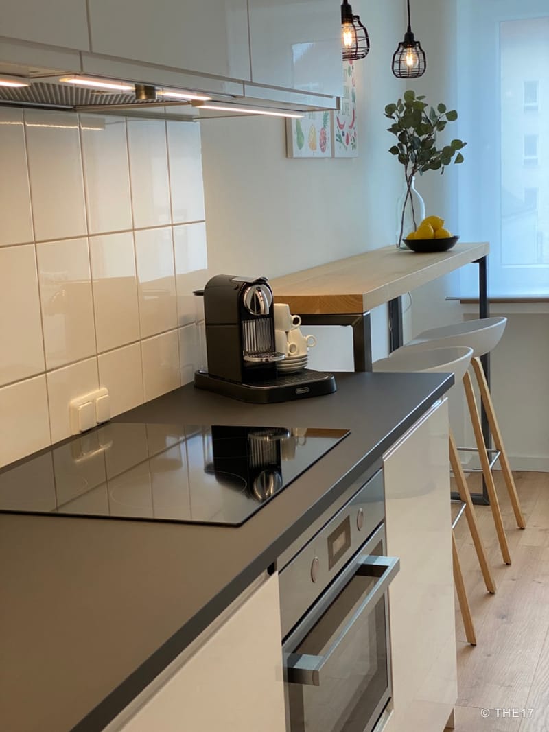 Moderne Küche mit Kochfeld, Kaffeemaschine, hellem Holz-Bar-Tisch, zwei weißen Barhockern und Fenster mit weißem Vorhang.