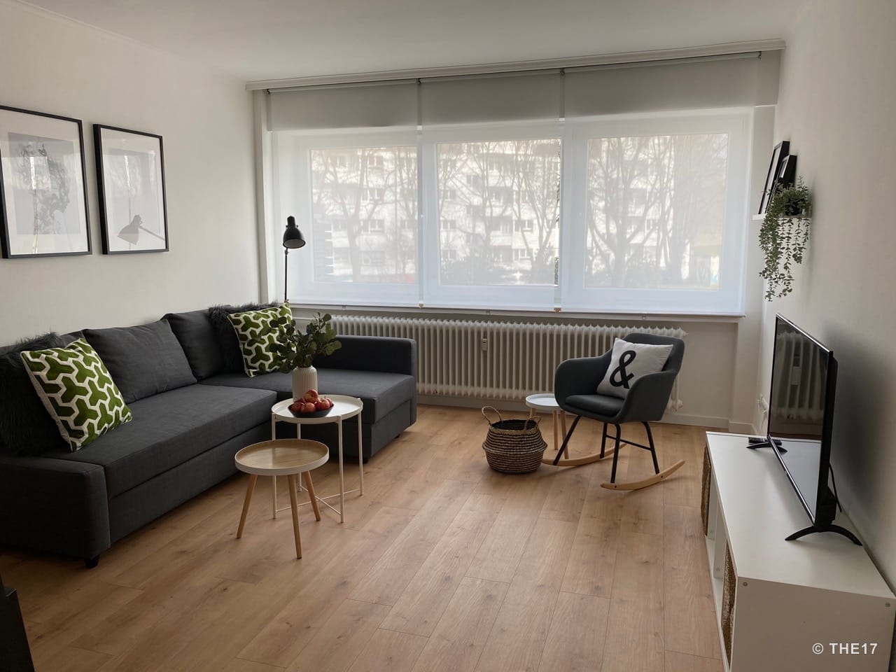 Helles Wohnzimmer mit grauem Ecksofa, grün-weißen Kissen, Schaukelstuhl, kleiner Tischgruppe und großem Fenster mit weißen Rollos.