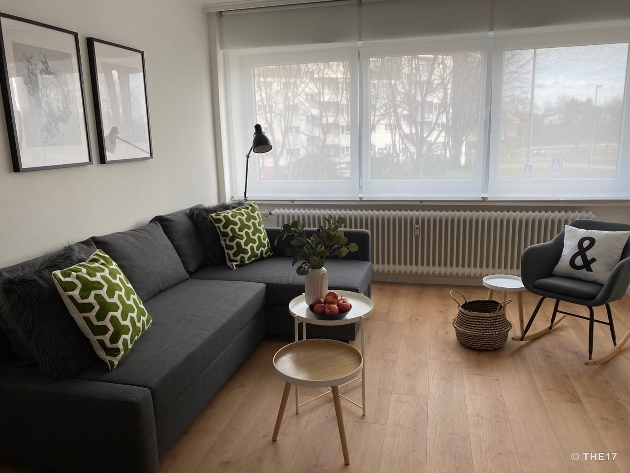 Helles Wohnzimmer mit grauem Ecksofa, grünen und schwarzen Kissen, Schaukelstuhl, Holz- und Metalltischen, großer Fensterfront mit Rollos.