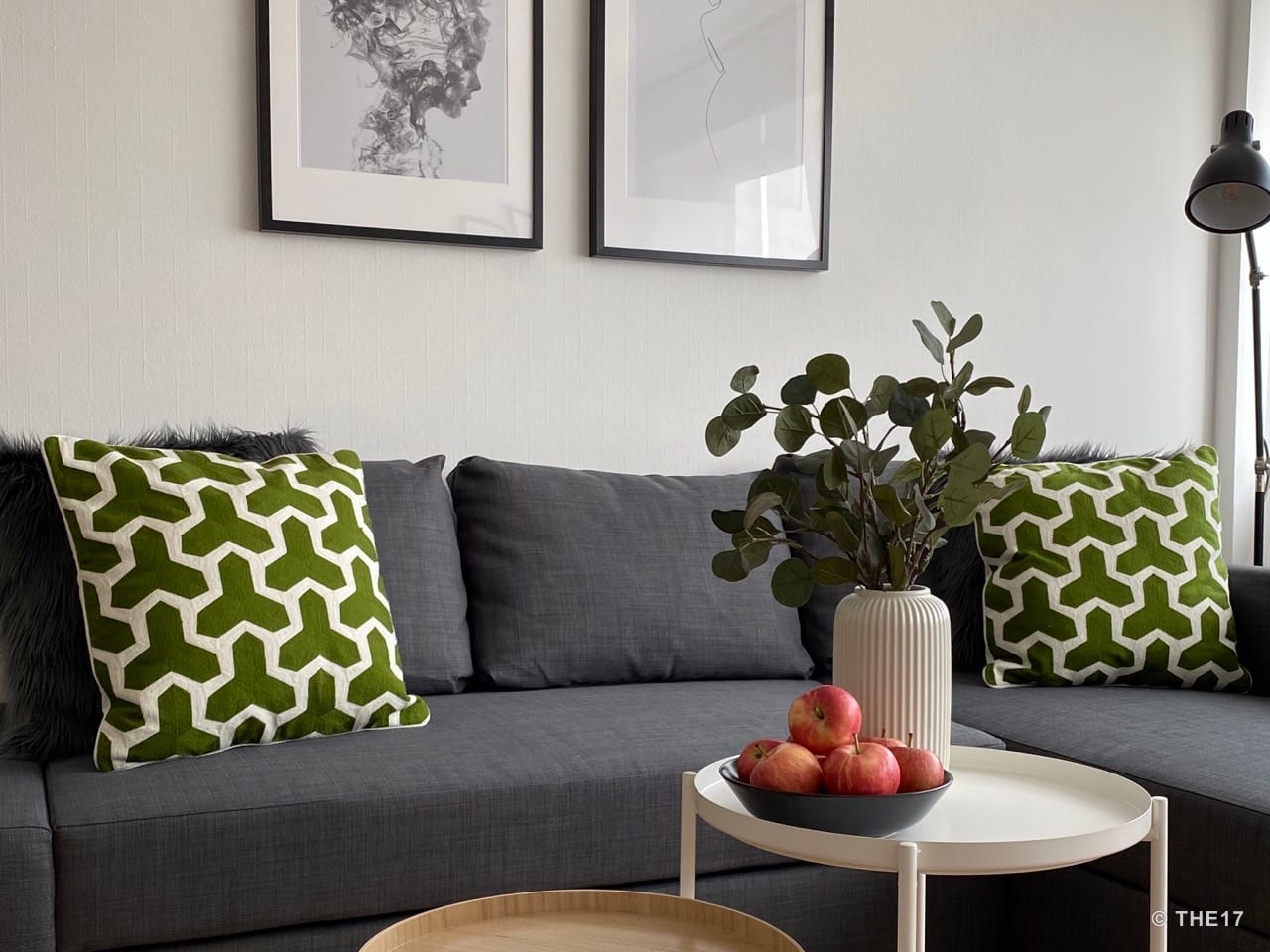 Graues Ecksofa mit grünen Kissen, schwarzem Kunstfell, zwei Wandbildern und einem weißen Beistelltisch mit Äpfeln und Pflanze.