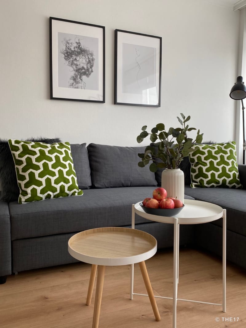 Hell eingerichtetes Wohnzimmer mit dunkelgrauem Sofa, zwei grünen Kissen, Holzboden und zwei Couchtischen, einer mit Vase und Obst.