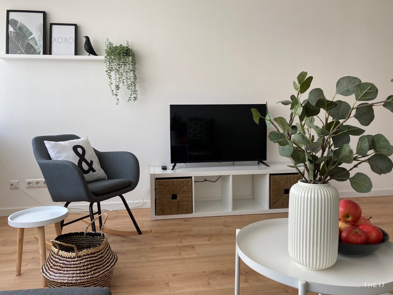 Wohnzimmer mit grauem Sessel, weißem Couchtisch mit Vase und Äpfeln, TV auf weißem TV-Board, Bilderrahmen und Pflanzen an der Wandregal.