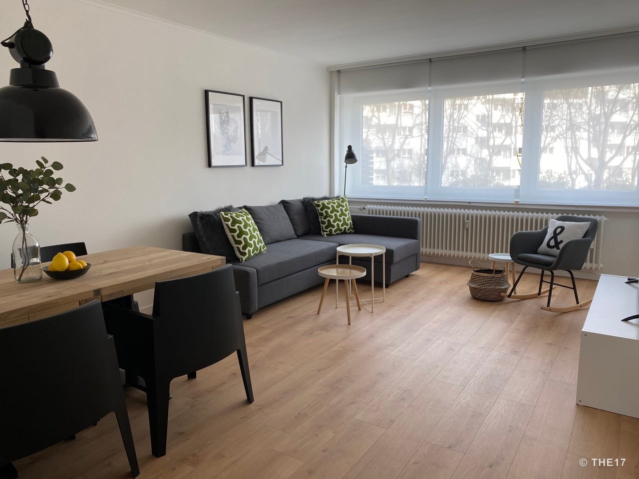 Helles Wohnzimmer mit grauem Ecksofa, zwei grünen Kissen, zwei Couchtischen und einem Essbereich mit Holztisch und schwarzen Stühlen.