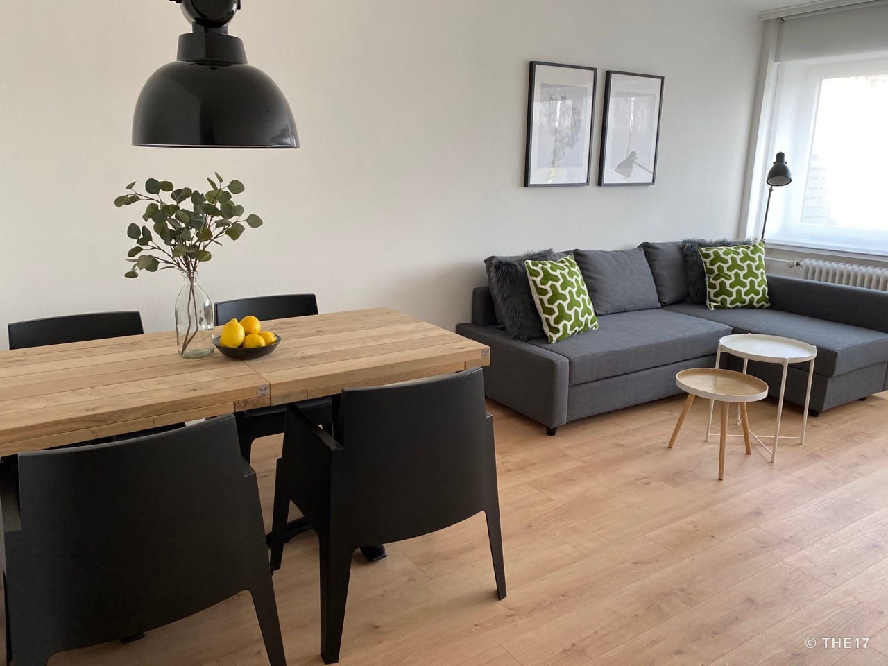 Wohnzimmer mit grauem Ecksofa, grün gemusterten Kissen, Holztisch mit schwarzen Stühlen und einer schwarzen Pendelleuchte.