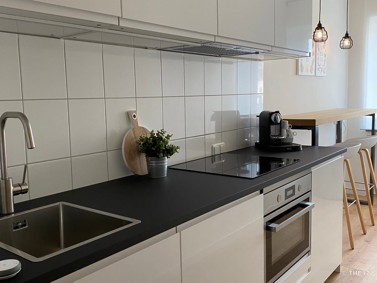 Moderne Küche mit schwarzer Arbeitsplatte, Edelstahlspüle, Cerankochfeld, Kaffeemaschine und Bar mit Hockern.