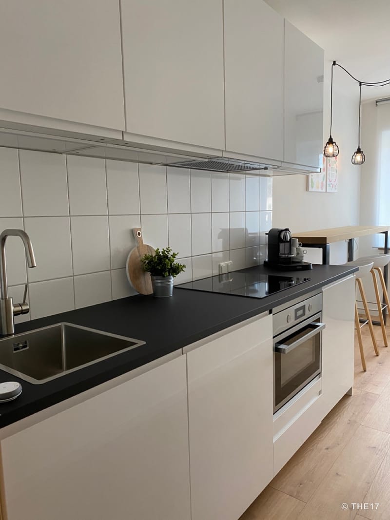 Moderne weiße Küche mit schwarzer Arbeitsplatte, Herd, Spüle, Kaffeemaschine und Barhockern am Fenster.