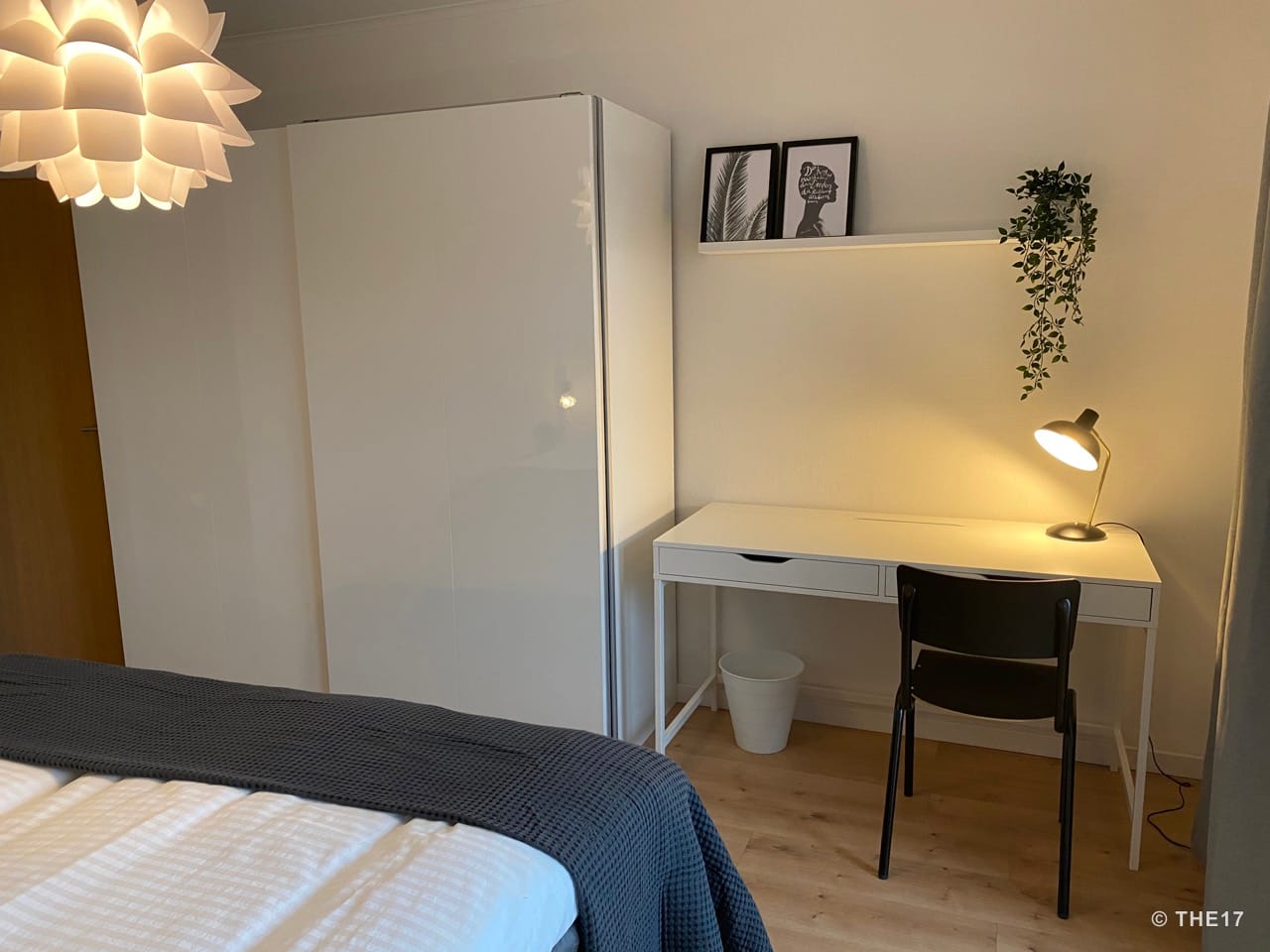 Schlafzimmer mit weißem Kleiderschrank, Schreibtisch, schwarzem Stuhl, Tischlampe und Wandregal mit Pflanzen und Bildern.