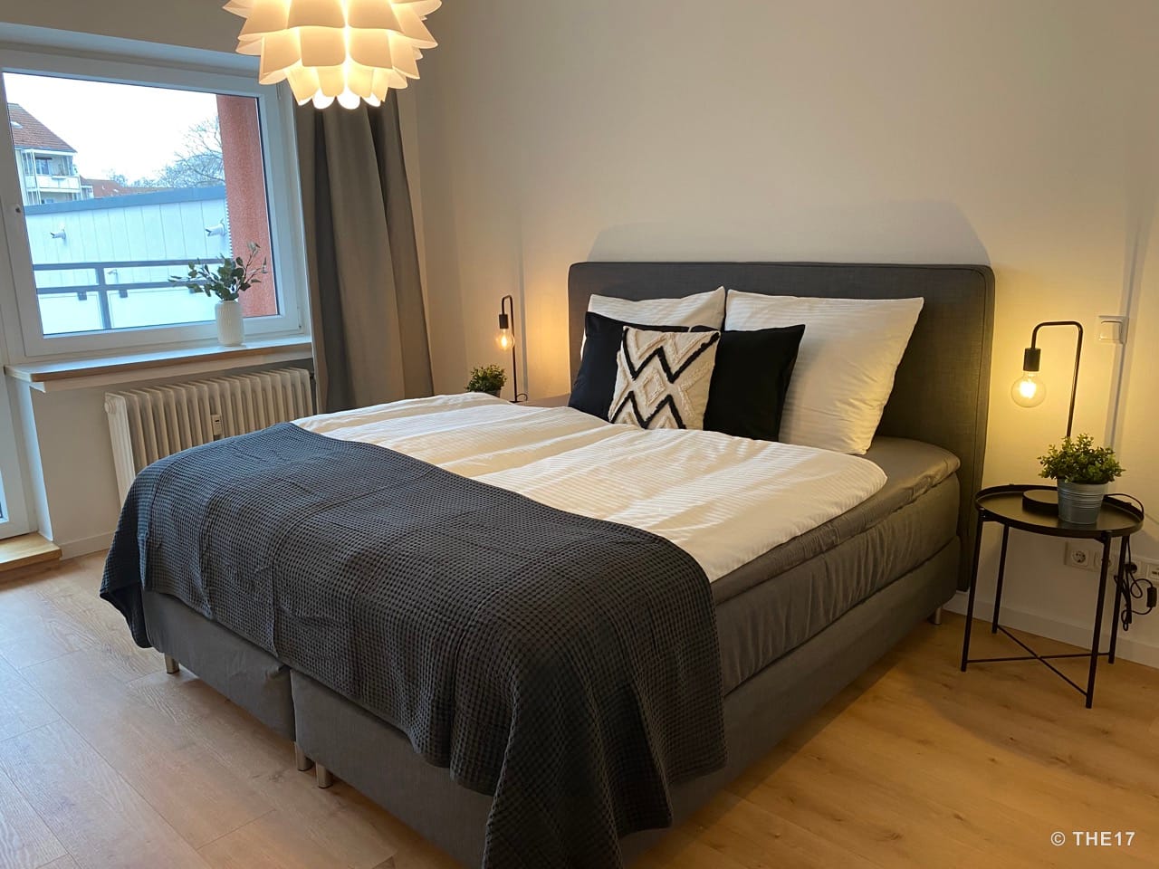 Modernes Schlafzimmer mit großem grauem Bett, weißen und schwarzen Kissen, seitlichem Nachttisch und warmem Licht.