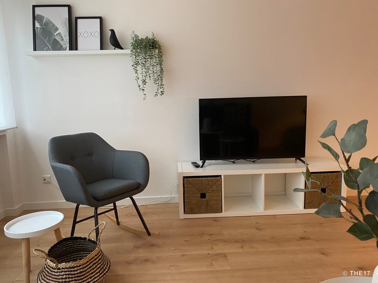 Wohnzimmer mit grauem Sessel, kleinem weißen Beistelltisch, TV auf weißem Lowboard und Wandregal mit Bilderrahmen und Pflanze.